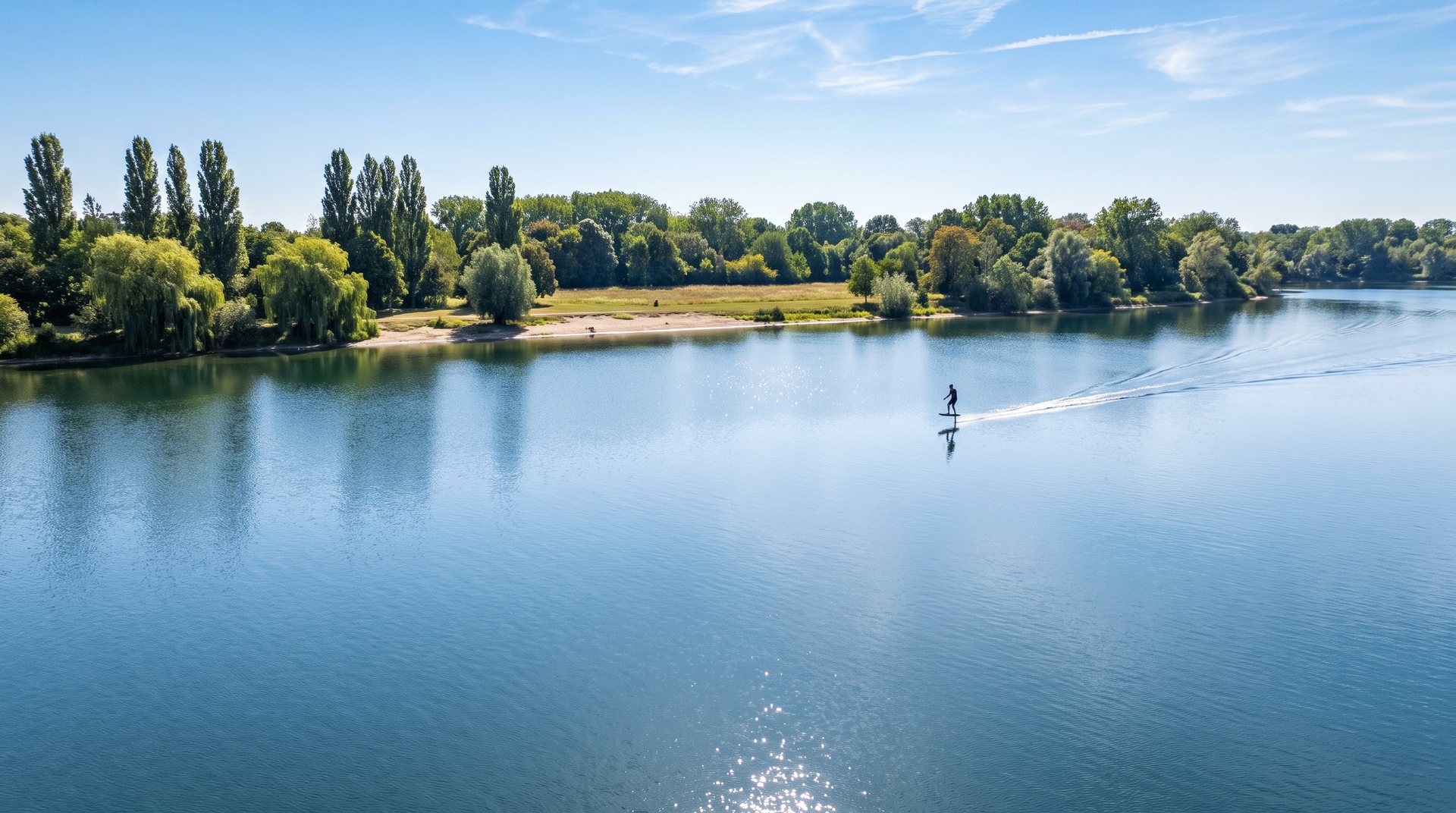 Foiler gleitet an einem sonnigen Tag über den Fühlinger See – Ufer mit Pappeln, Weiden und Wiese